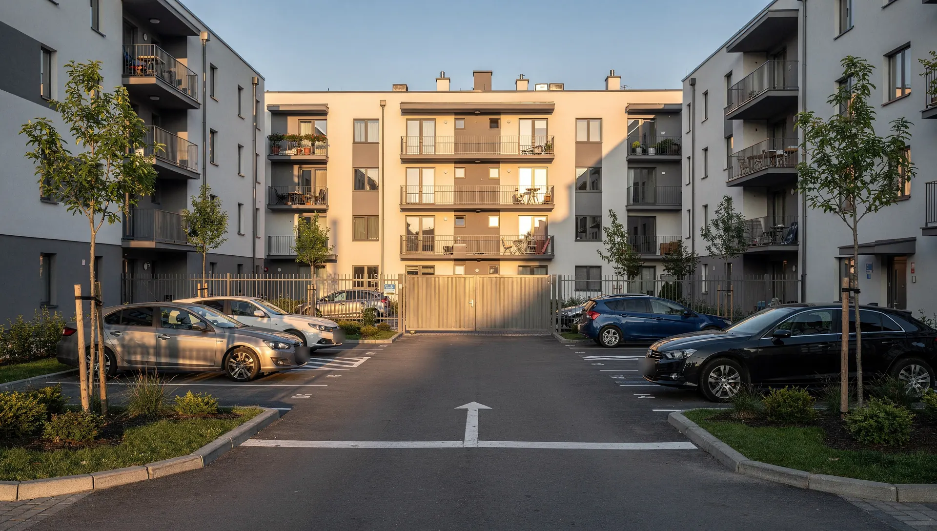 Parking na dziedzińcu osiedla z wyznaczonymi miejscami i bramą w ogrodzeniu.
